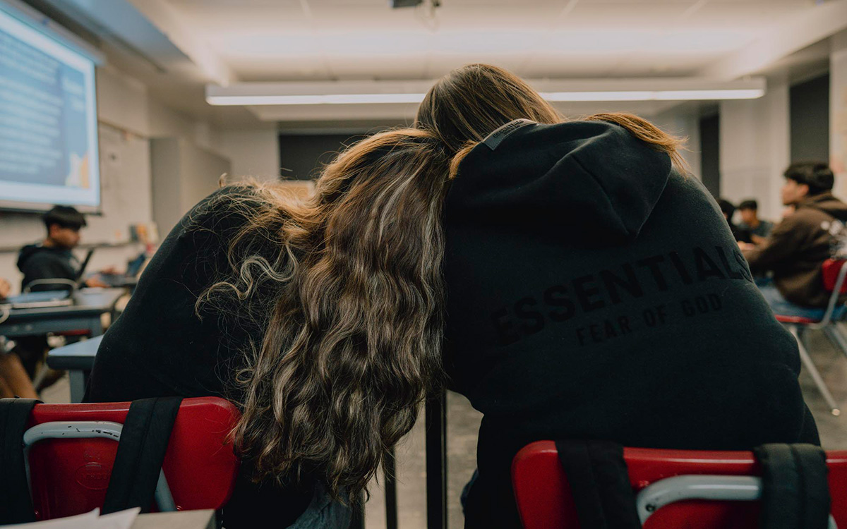 Photo taken from behind two high school students with long hair sitting at desks in a classroom, leaning against each other
